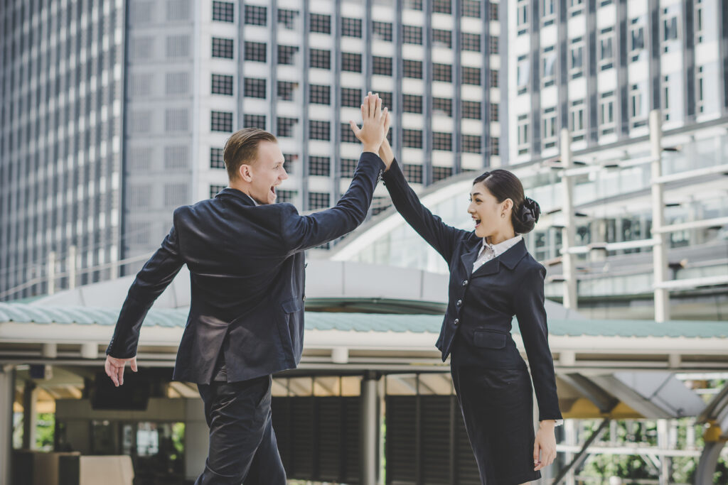 Two business professionals celebrating success with a high-five outside a modern office building as they have successfully finish this month's task with the use of payroll automation.