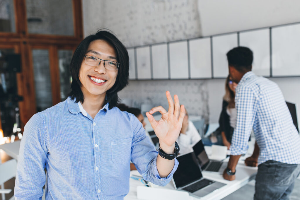 Happy Asian man showing an okay sign in an office, reflecting employee satisfaction with well-managed leave.