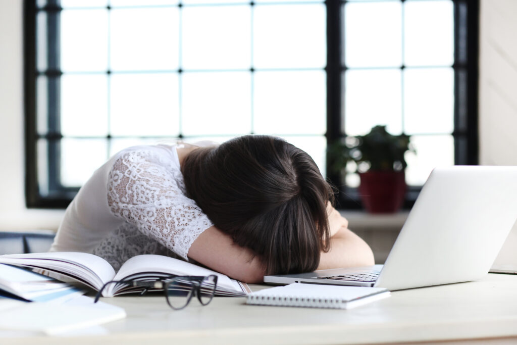 Tired woman with head down on desk, overwhelmed with work, next to a laptop.