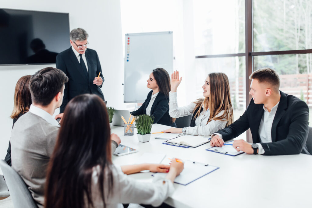 A diverse team in a meeting, with one employee raising a hand to provide feedback on the attendance management system.