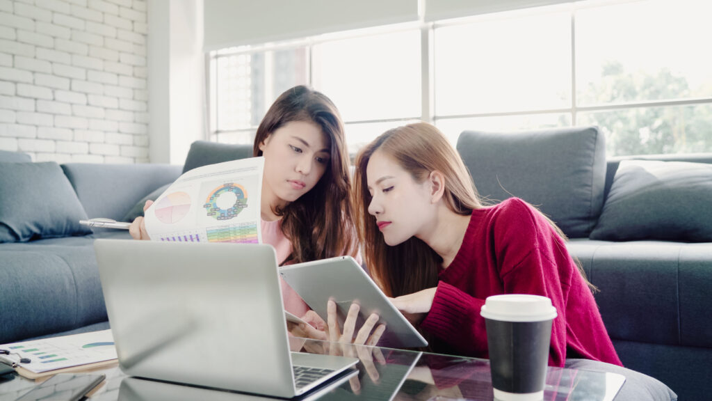 Two businesswomen comparing software options on a tablet and printed chart, discussing features for attendance management.