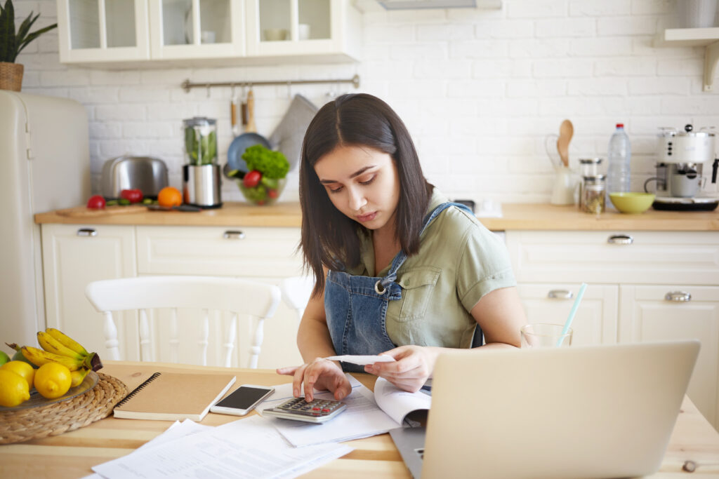 A young woman calculating utility bills at a kitchen table with documents, a calculator, and a laptop.