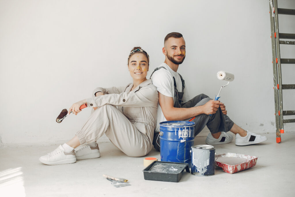 A young couple sitting with paint rollers and buckets, smiling during a home renovation project.