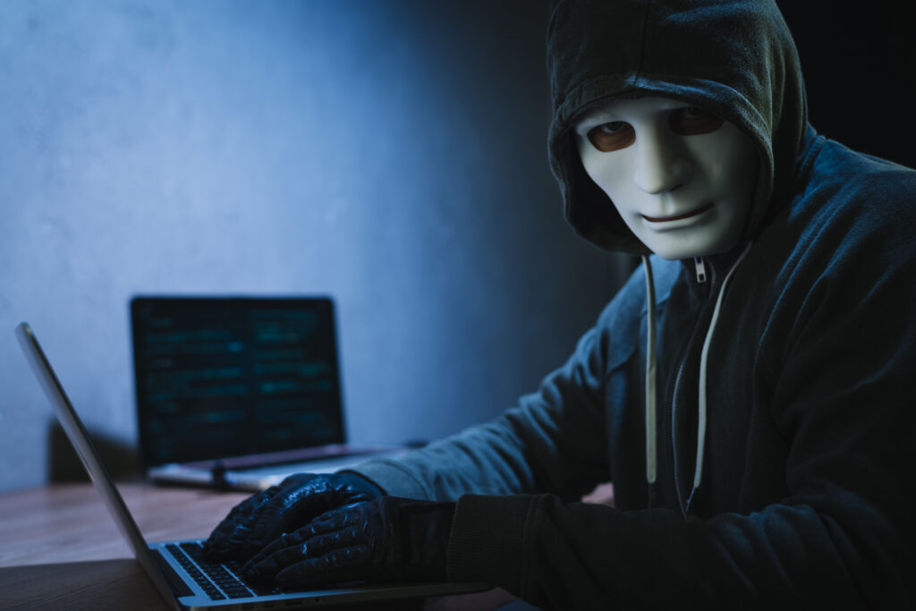 A masked hacker wearing gloves using a laptop in a dark room, symbolizing cybersecurity threats.