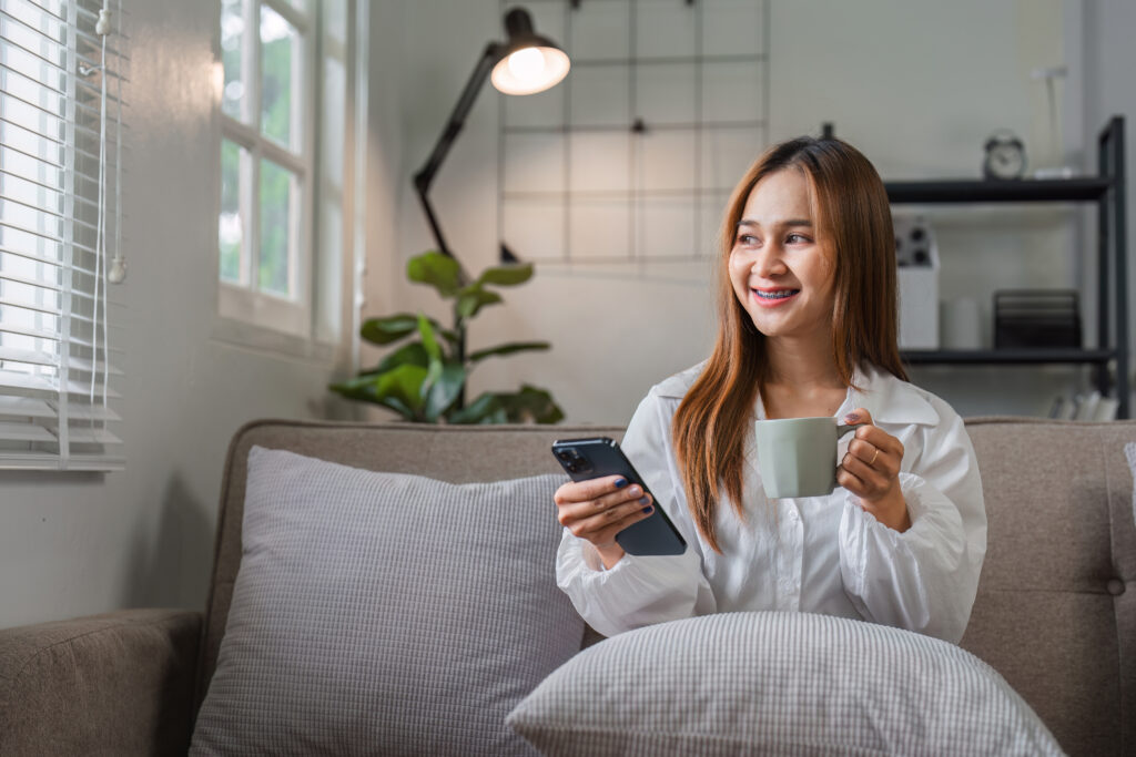 A smiling woman relaxing with coffee while using Facility Booking Technology on her smartphone at home.